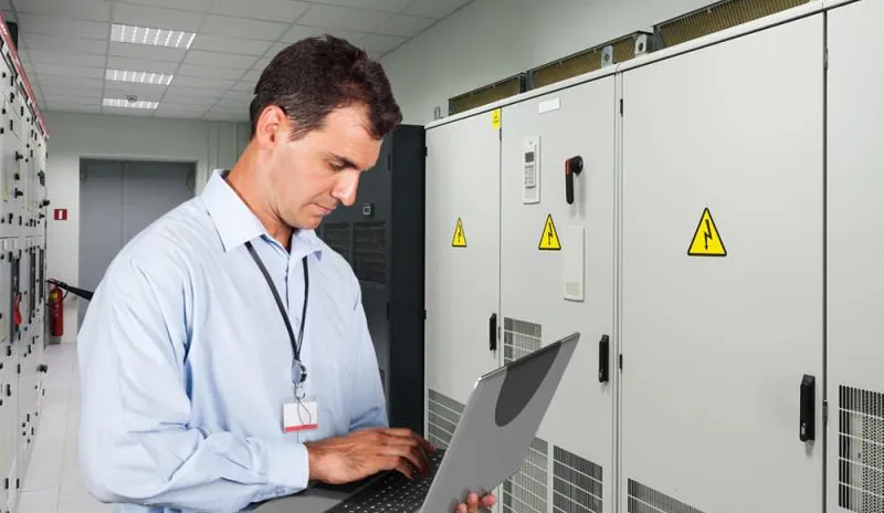 Technical professional reviewing data on a laptop beside electrical equipment